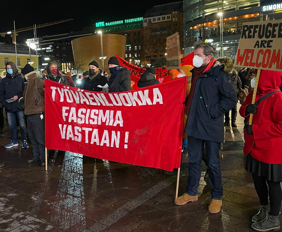Mielenosoittajia seisoo öisellä mutta valaistulla torilla. Kaksi henkilöä pitelevät suurta punaista banderollia, jossa lukee "Työväenluokka fasismia vastaan!". Näkyvissä on myös kyltti, jossa lukee "Refugees are welcome".